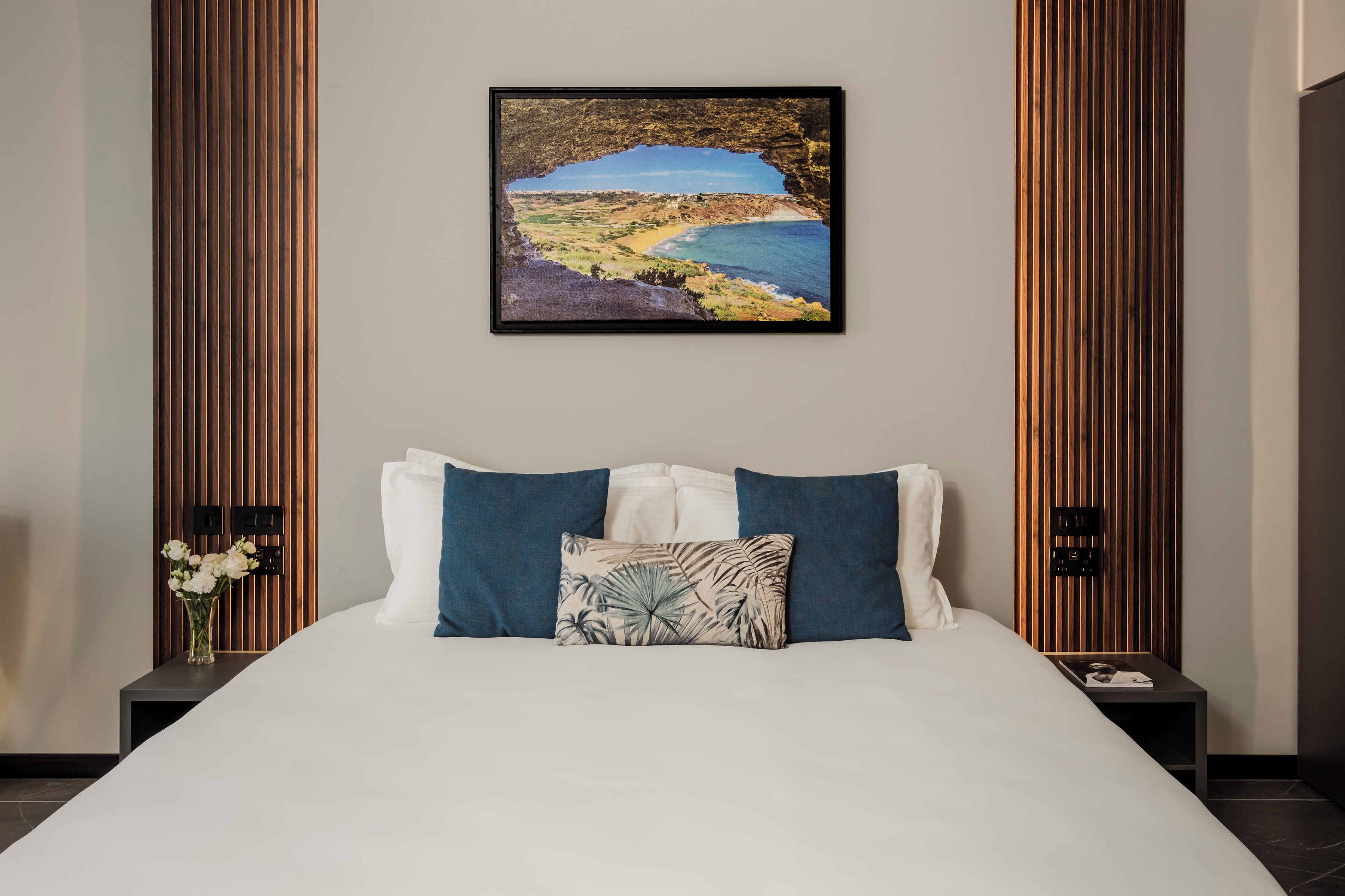 A neatly made bed with a white duvet and two pillows, a framed coastal landscape painting above it, and a nightstand with a vase of flowers to the left.