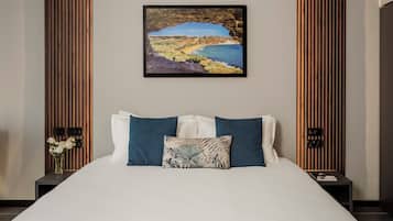 A neatly made bed with a white duvet and two pillows, a framed coastal landscape painting above it, and a nightstand with a vase of flowers to the left.