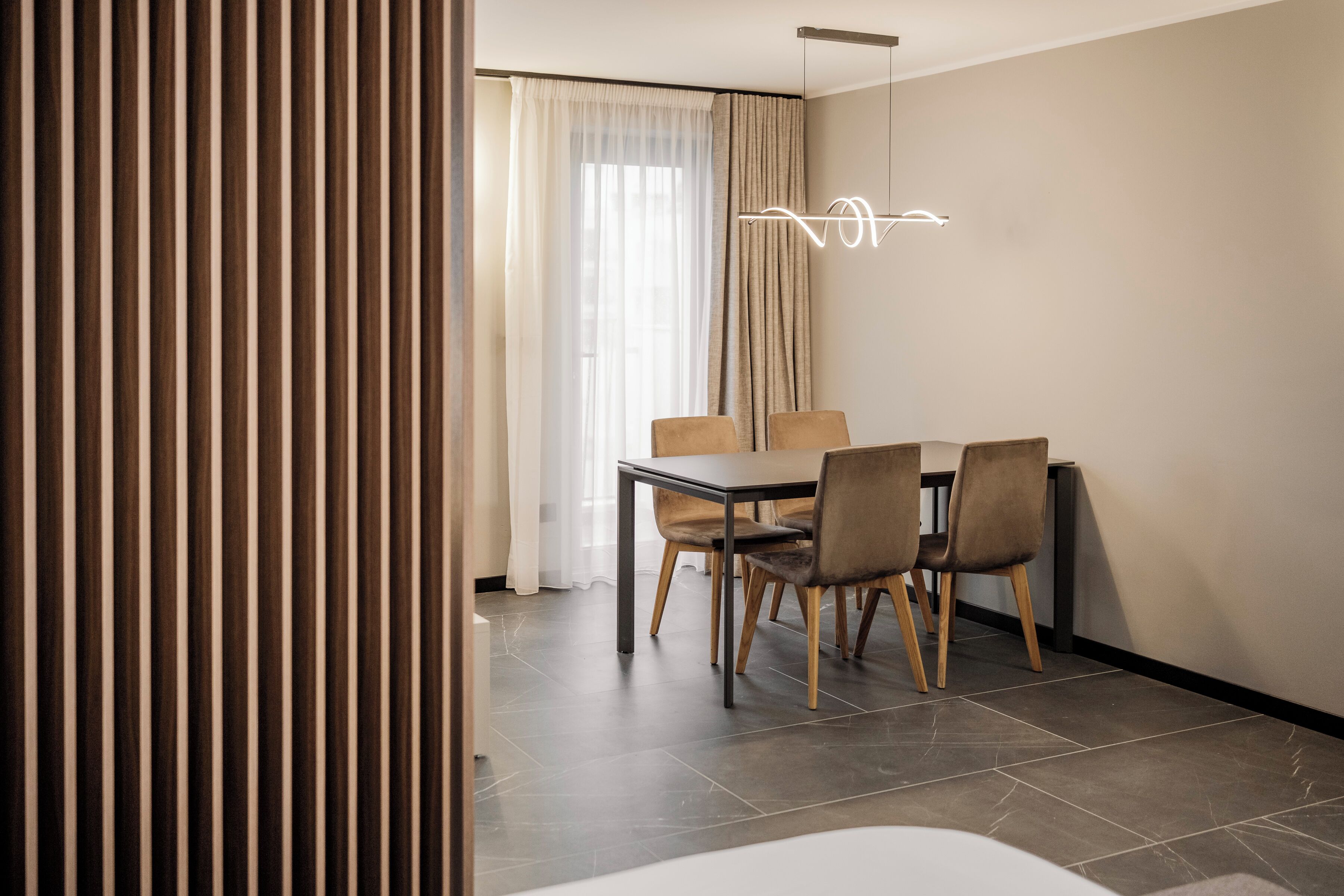 A modern dining area with a table and chairs, a light fixture, and a view of the outdoors through a window with curtains.