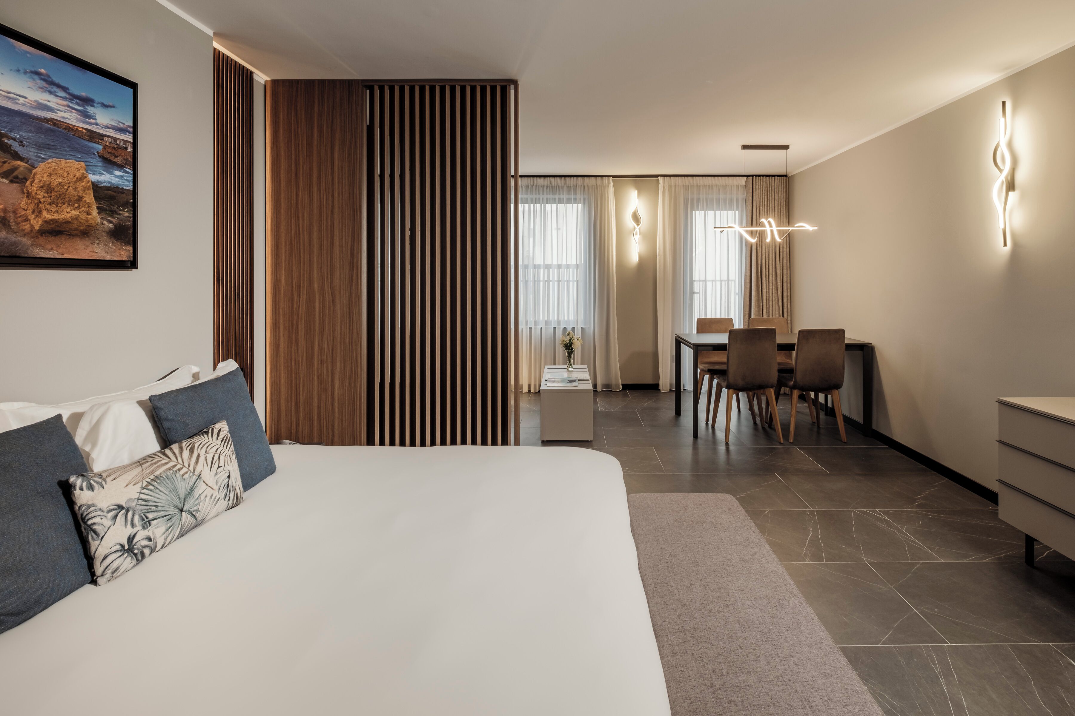 A modern hotel room with a large bed, a dining area with chairs, and a view of the sea through the window.