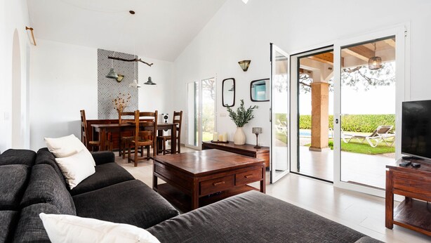 Un salon moderne avec un canapé foncé, une table basse en bois et un meuble pour la télévision. On y trouve également un coin repas avec une table et des chaises, ainsi qu’une grande fenêtre donnant sur l’extérieur.