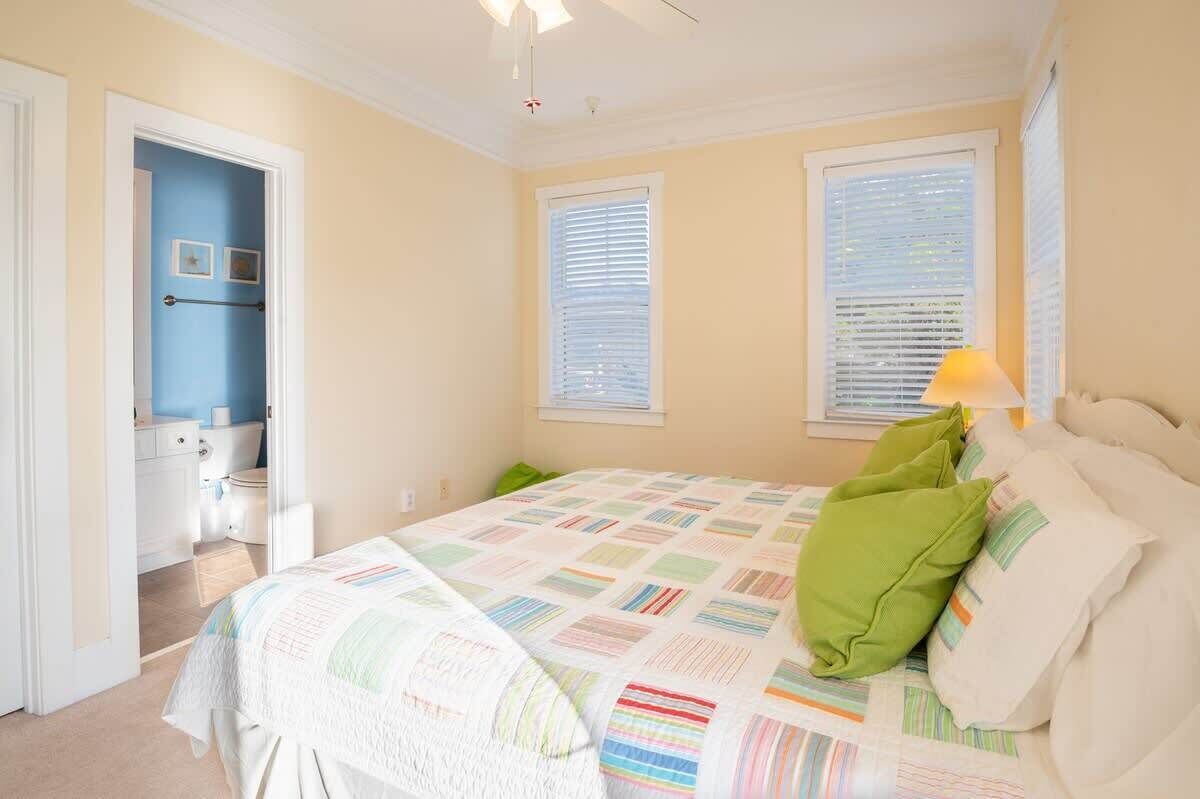 Un dormitorio con cama, dos ventanas con persianas, una lámpara de noche y una puerta que da a un baño.