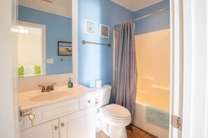 Une salle de bain avec un lavabo, des toilettes et une baignoire, tous de couleur blanche. Deux tableaux encadrés ornent le mur, ainsi qu’un porte-serviettes et un rideau de douche.