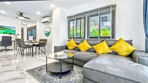 Uma sala de estar moderna com um sofá cinzento, almofadas amarelas, uma mesa de centro de vidro e uma área de jantar com cadeiras e uma mesa.
