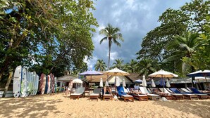 Uma praia com cadeiras de praia e guarda-sóis, um suporte para pranchas de surf e palmeiras.