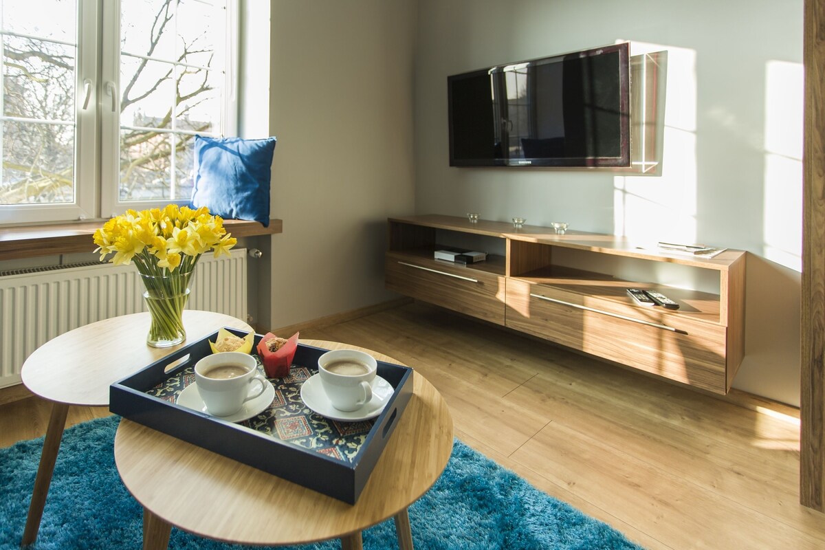 Een moderne woonkamer met een houten vloer, een blauw vloerkleed, een bijzettafeltje met een dienblad vol koffiekopjes en gebak, een vaas met gele bloemen, een blauw kussen en een aan de muur bevestigde televisie.