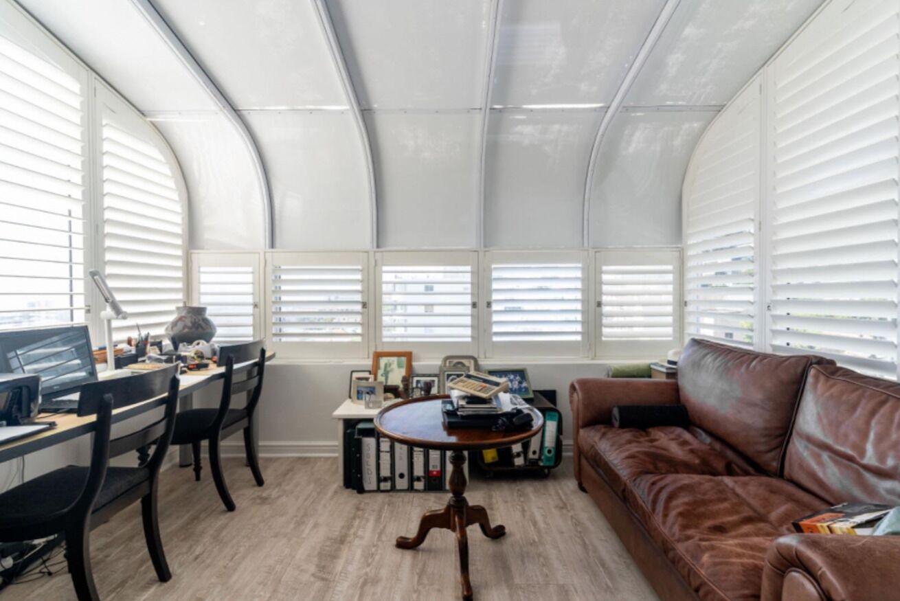 Una habitación con techo curvo, un escritorio con ordenador, un sofá de cuero marrón y una pequeña mesa redonda con libros y una lámpara.