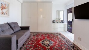 Ein Zimmer mit grauem Sofa, einem Teppich mit roten und schwarzen Mustern, einem weißen Kleiderschrank, einem Deckenventilator und einem flachbildfernseher an der Wand.