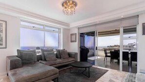A spacious living area with a sectional sofa, a coffee table, and large windows offering a view of the city. - Cozy 2 Bedroom Beachfront Apartment (Cape Town)