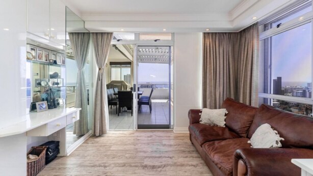 A modern living room with a brown leather sofa, a glass cabinet with decorative items, and a view of the city through large windows. - Cozy 2 Bedroom Beachfront Apartment (Cape Town)