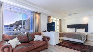 A modern hotel room with a leather sofa, a bed with a dark headboard, a flat-screen TV, and a view of a cityscape through large windows. - Cozy 2 Bedroom Beachfront Apartment (Cape Town)