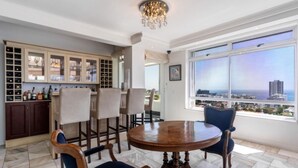 A dining area with a round wooden table and chairs, a chandelier, and a view of the city through large windows. - Cozy 2 Bedroom Beachfront Apartment (Cape Town)