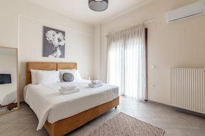 A bedroom with a bed, a bedside table, a mirror, a window with curtains, and an air conditioning unit. - Zephyrus Cityscape Condo, Apartment (Heraklion)