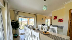 A dining area with a long table, white chairs, a potted plant, and a view through the glass doors. - Villa Nosredna 5 Bedroom & Pool LES01 (Ayamonte)