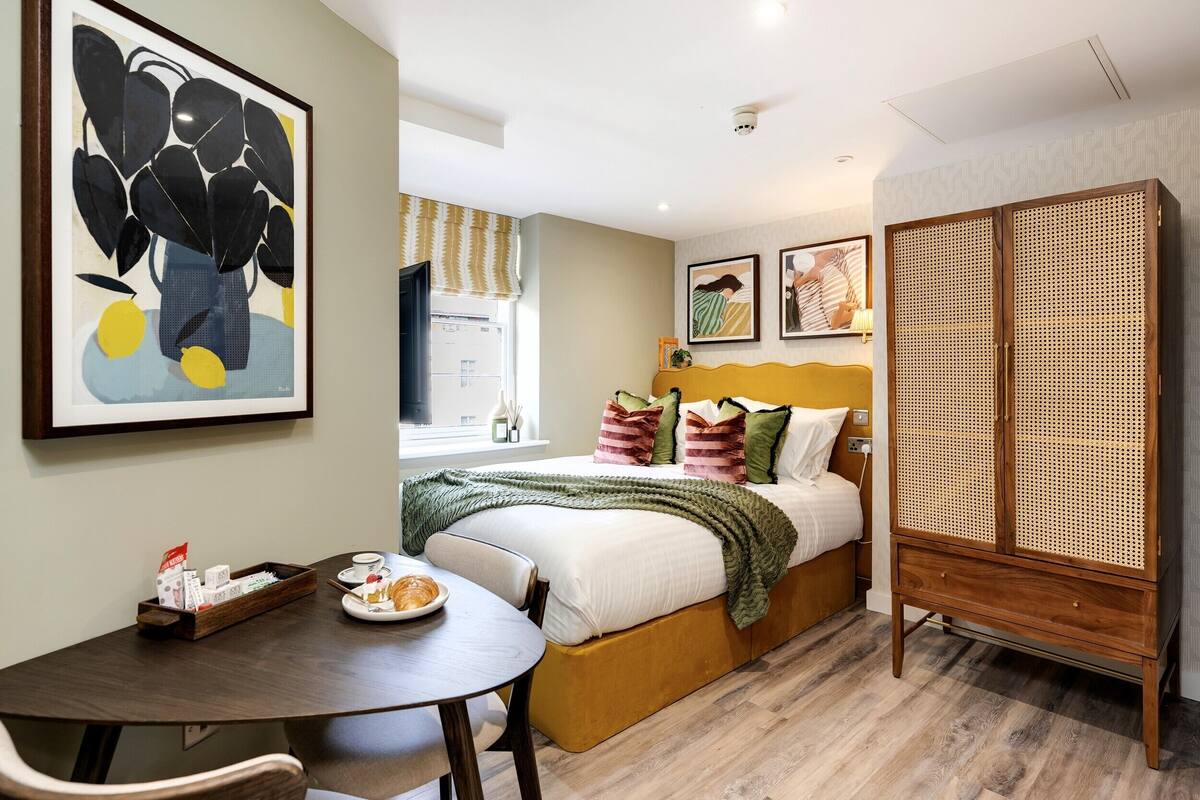 Quarto de hotel com cama, guarda-roupa de madeira, mesa de jantar e prato de comida, e um quadro na parede.