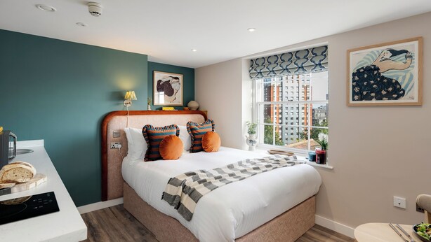 Une chambre moderne avec un grand lit, une table de chevet avec une lampe et une vue sur la ville par la fenêtre.