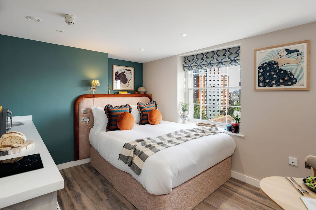Quarto moderno com uma cama grande, uma mesinha de cabeceira com luminária e vista para a cidade pela janela.
