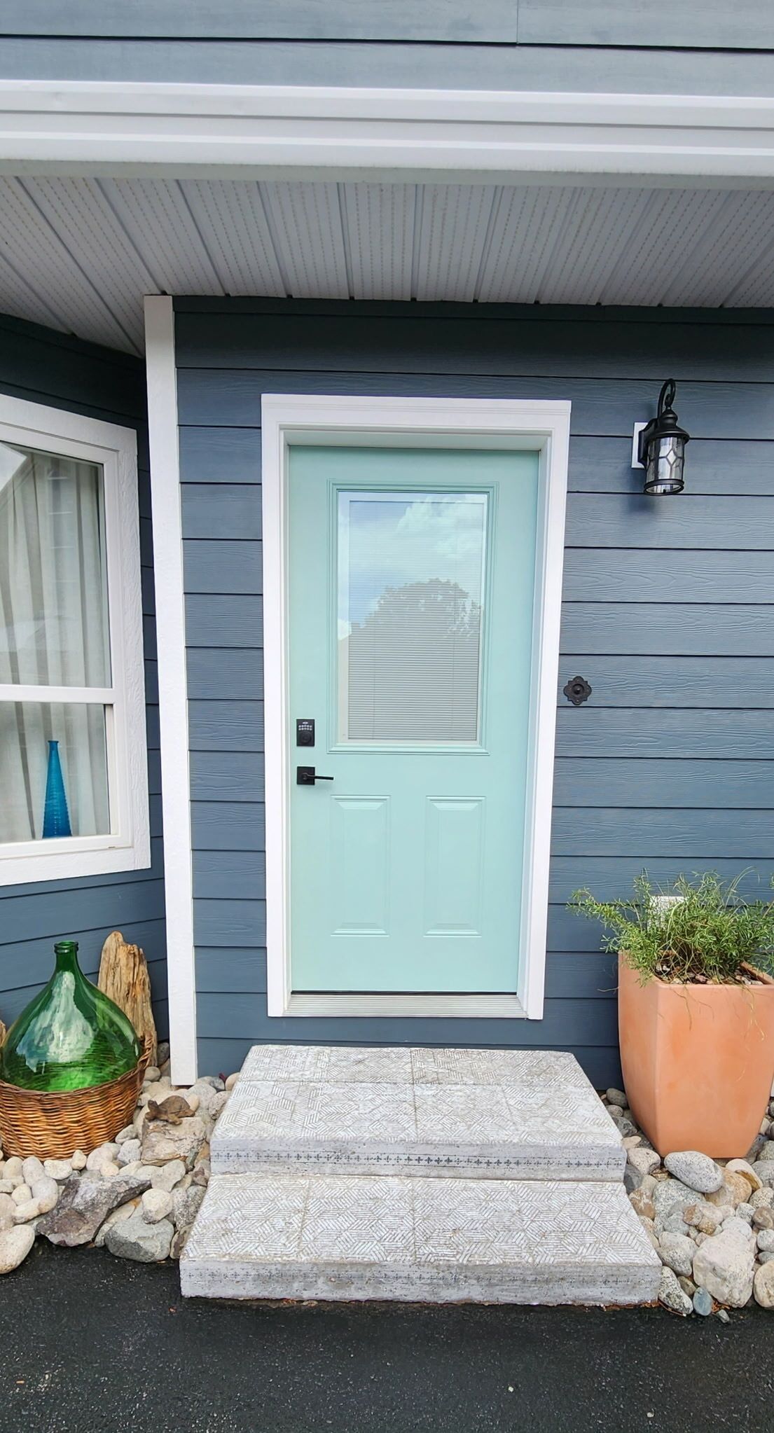 Ein Haus mit einer hellblauen Tür, einer kleinen Veranda mit Fußmatte, einem Blumentopf und einem Fenster mit Vorhang.