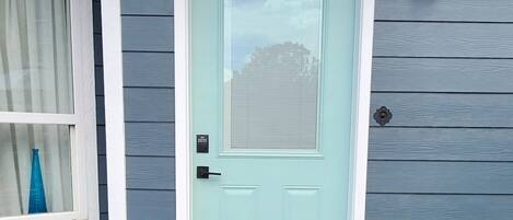 A house with a light blue door, a small porch with a welcome mat, a potted plant, and a window with a curtain.