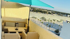 Un balcón con muebles de mimbre, un paraguas color turquesa y vista a un edificio moderno y palmeras.