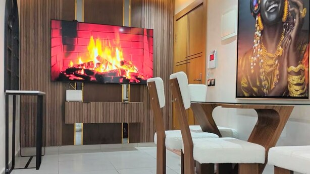 Un comedor moderno con un televisor de pantalla plana grande mostrando una chimenea, sillas de madera y un cuadro grande en la pared.