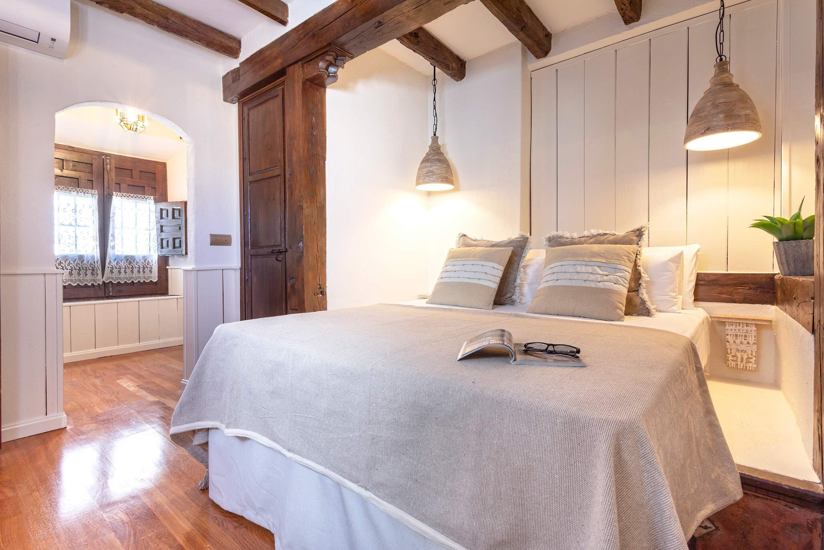A bedroom with a bed, wooden beams, a hanging light, and a window with curtains.