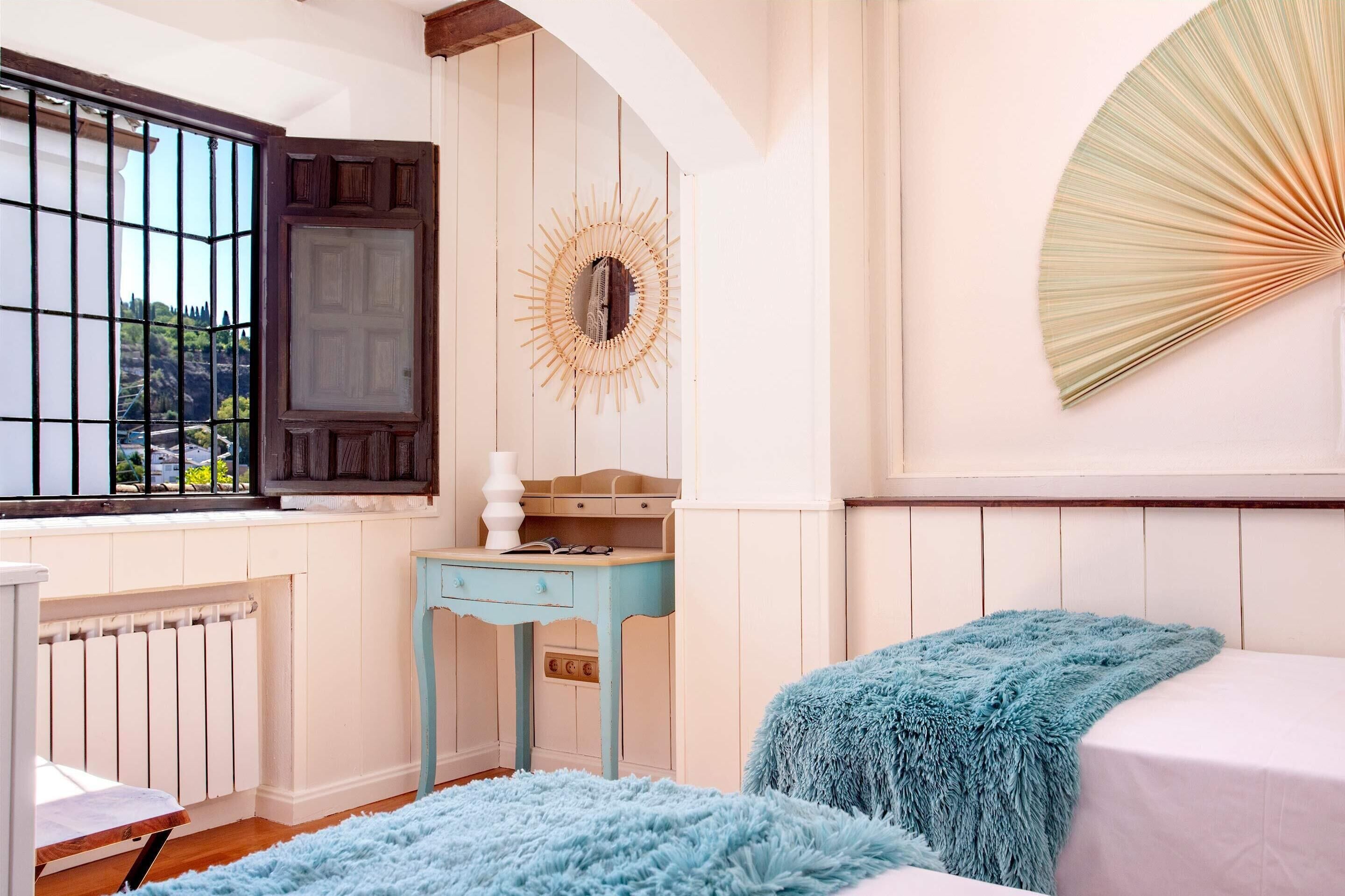 Ein Schlafzimmer mit Bett, Kommode, einem Fenster mit Gitter und Blick ins Freie.