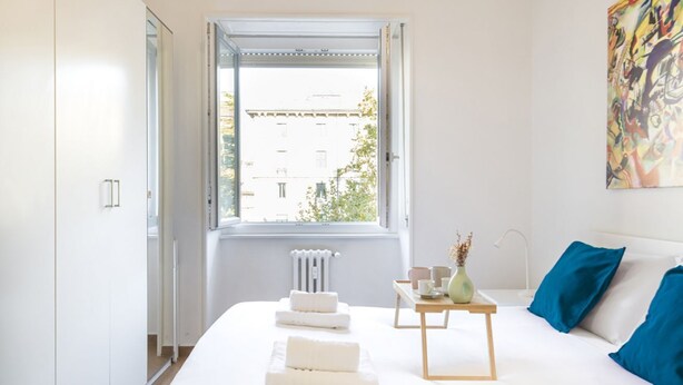 Un dormitorio moderno con un ventanal grande, una cama con sábanas blancas, una mesita de noche con un jarrón y un cojín azul.