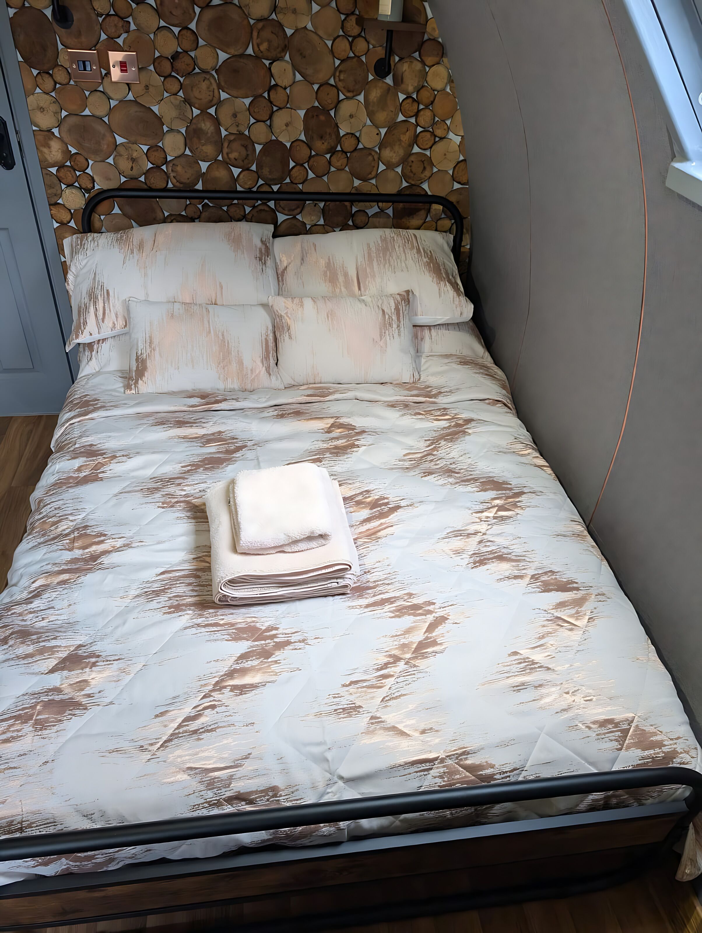 Una cama con un edredón blanco y marrón estampado, una toalla blanca doblada cuidadosamente y un cabecero con diseño de tronco de madera.