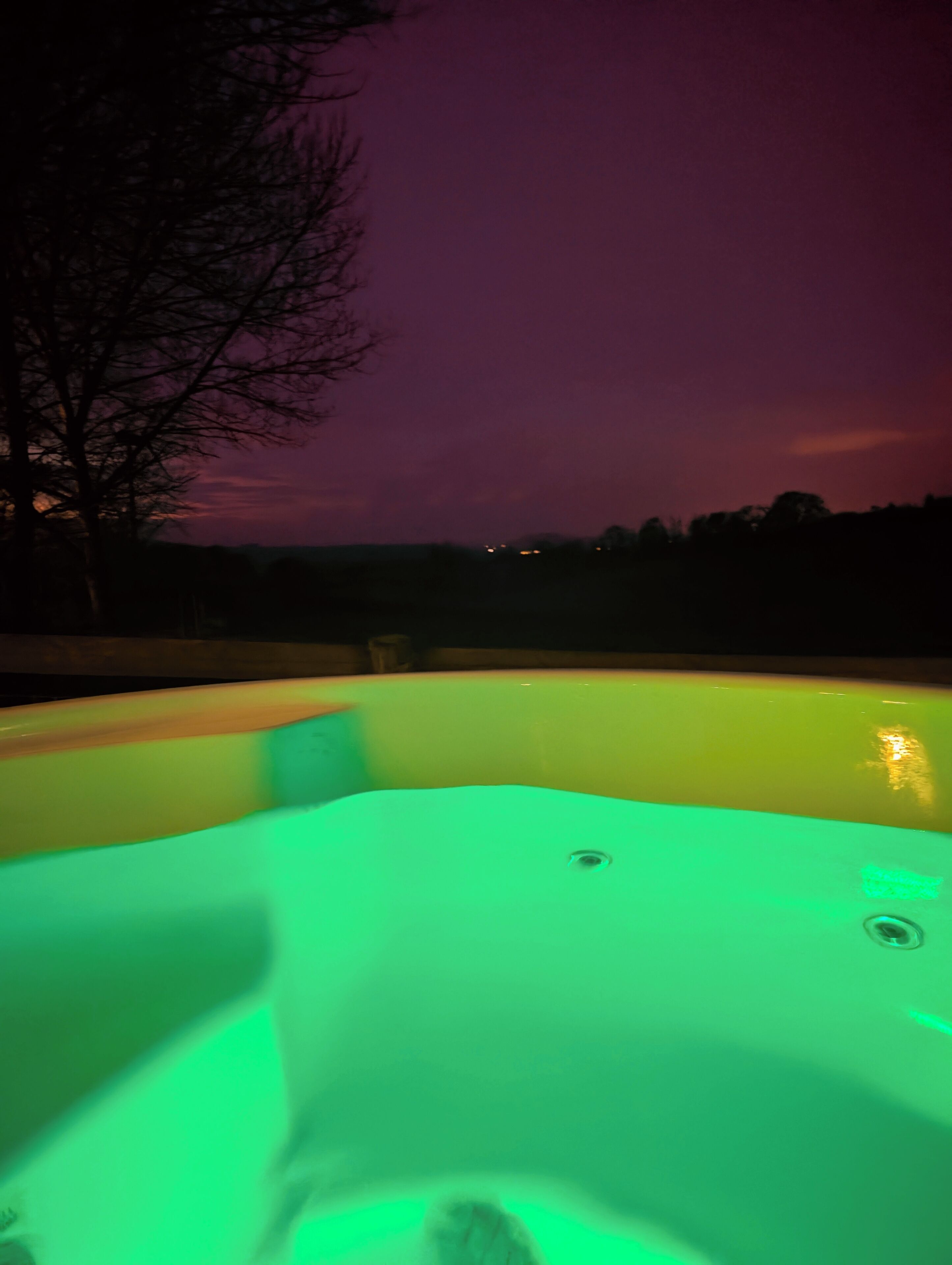 A hot tub with a green light, surrounded by a dark landscape and a purple sky.