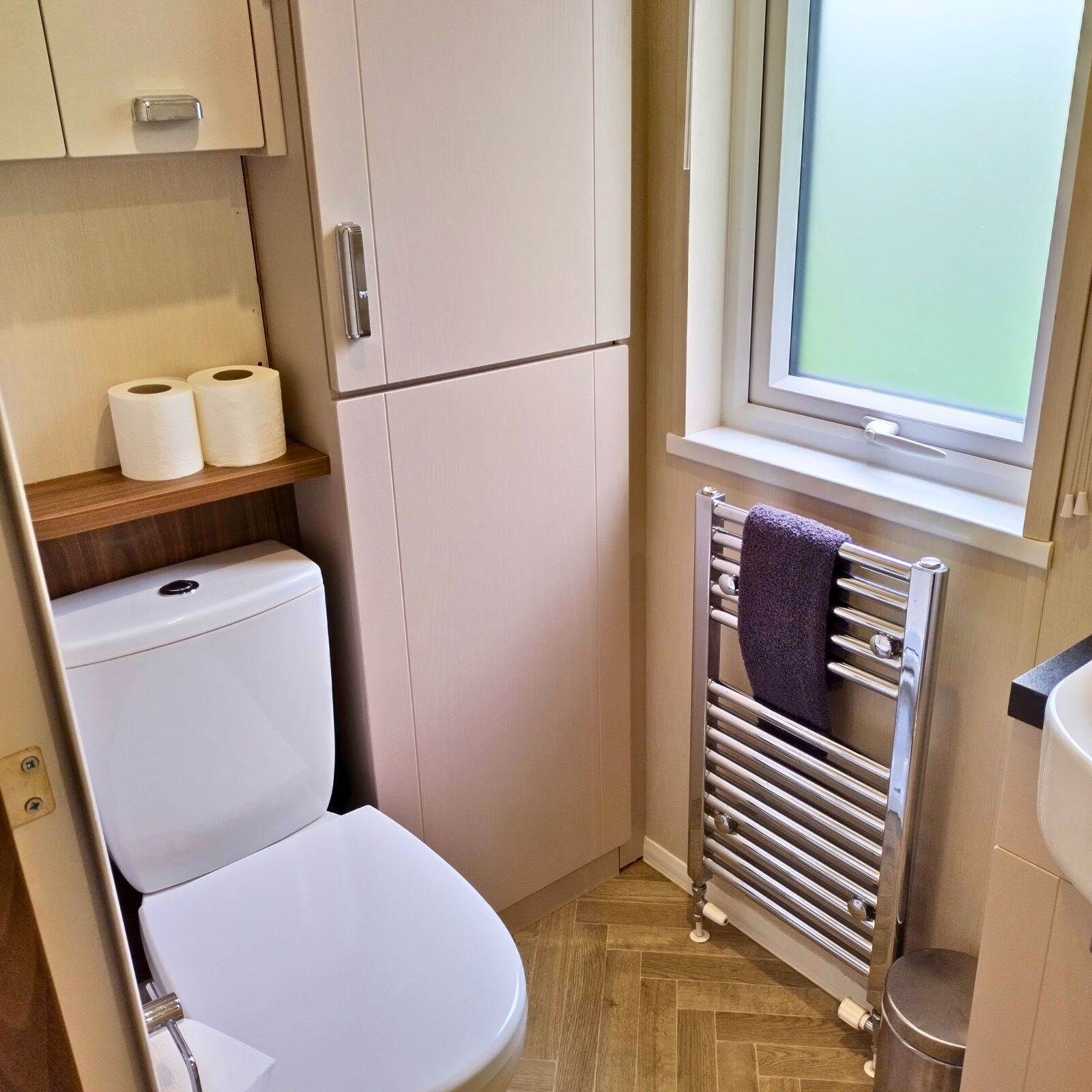 A compact bathroom with a toilet, a shelf with toilet paper, a towel rack with a towel, and a window with a view.