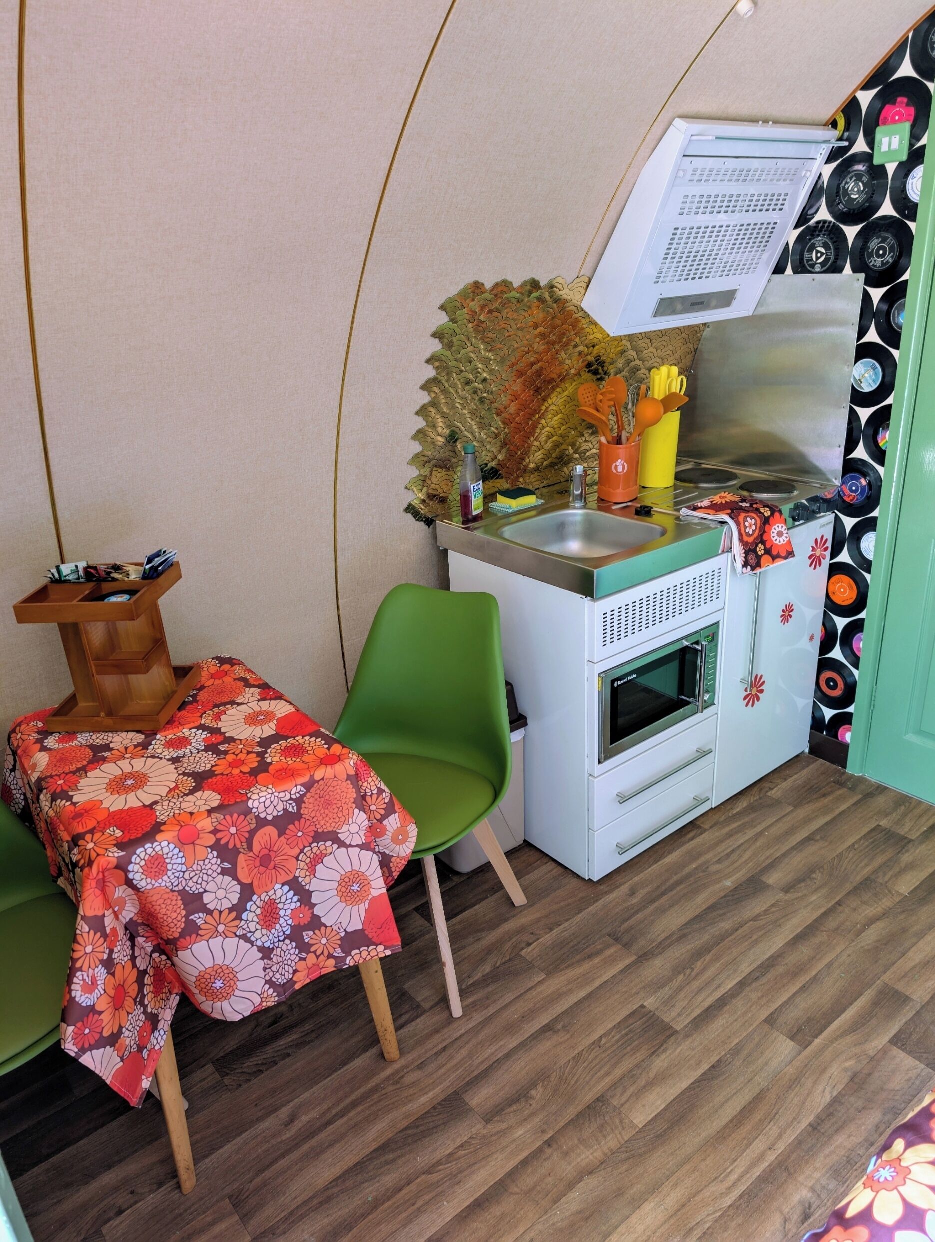 Una kitchenette compacta con un lavabo, estufa y armarios de almacenamiento, una pequeña mesa de comedor con mantel floreado y una silla verde.