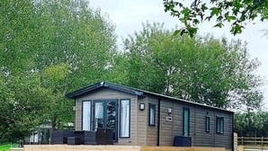 Een gelijkvloers, modern houten vakantiehuis met een plat dak en grote ramen. Het heeft een overdekte veranda met buitenmeubilair en een houten hek op de voorgrond.