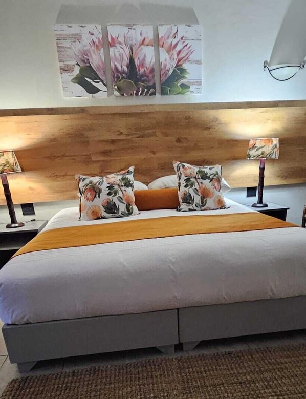 A bedroom with a wooden headboard, a large bed with floral pillows, a bedside table with a lamp, and a wall-mounted lamp. - Mont Aux Sources Drakensberg Resort (Jagersrust)
