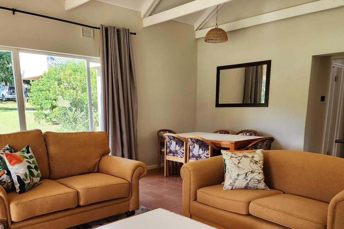 A living room with two sofas, a coffee table, a dining table with chairs, and a large window with curtains.