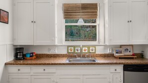 A kitchen with white cabinets, a granite countertop, a built-in sink, a window with bamboo blinds, and a hanging pendant light. - The Yellow Rose of SoCo:Charming Home Steps from South Congress and 1 mile to Downtown (Austin)
