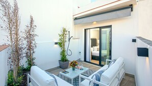 Un espace extérieur moderne, aménagé avec des meubles blancs, une plante en pot et offrant une vue sur l’intérieur à travers une porte vitrée.