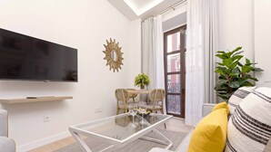 Un salon moderne avec une table basse en verre, un téléviseur à écran plat fixé au mur, un coin repas avec une table et des chaises, et une grande plante en pot.