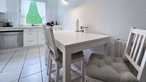 A kitchen with white cabinetry, a dining table, chairs, a hanging light fixture, and a view of greenery outside. - Alpha Apartments: Central 2BR, Living Room & Bath (Essen)
