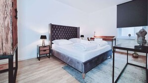 A modern bedroom with a tufted headboard, a bed with white linens, a bedside table with a lamp, a small table with a plant, and a large window.