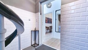 Un interior modern amb una paret de maó blanc, una escala, una taula petita amb un objecte decoratiu, i una zona de dinar visible a través d'una porta oberta.