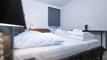 A hotel room with a bed, pillows, a bedside table, a lamp, a window, and a view of the sky.