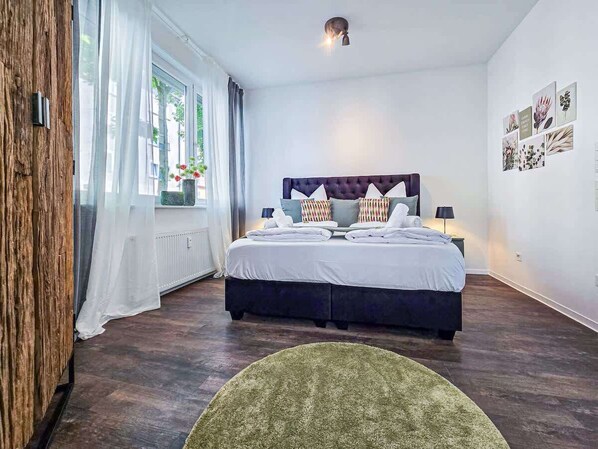 A bedroom with a large bed, a wooden wardrobe, a window with sheer curtains, and a wall with framed pictures.