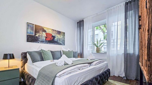 A bedroom with a large bed, a bedside table, a window with curtains, and a wall-mounted painting.