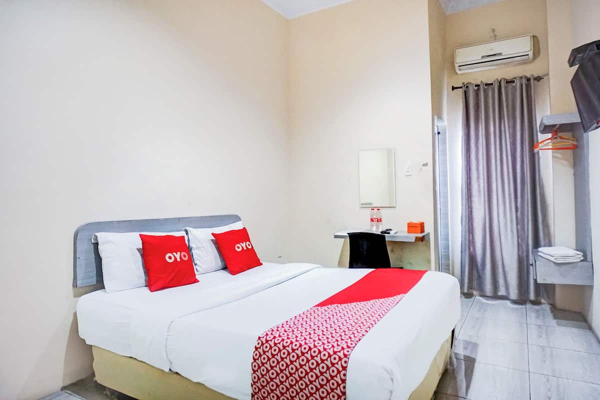Une chambre d’hôtel avec un lit, une couverture rouge et blanche, un oreiller rouge, un oreiller blanc, une tête de lit grise, un climatiseur fixé au mur, un rideau, une petite table avec une bouteille et un miroir.