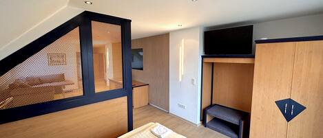 A modern hotel room with a wooden floor, a bed with a white mattress, a small seating area, a TV mounted on the wall, and a wooden wardrobe.