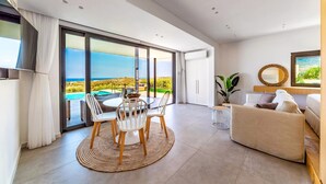 Una sala de estar moderna con un gran ventanal corredero que da a un balcón con vistas al océano. Hay una mesa de comedor redonda con sillas, un sofá y un televisor montado en la pared.