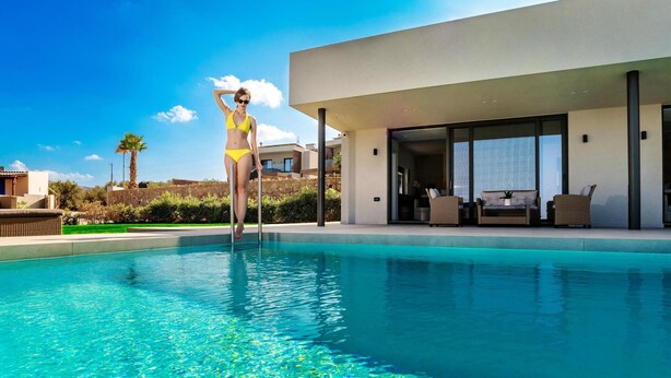 Un área moderna junto a la piscina con una pileta azul cristalina, una persona en traje de baño amarillo y un edificio contemporáneo con amplios ventanales y mobiliario exterior.