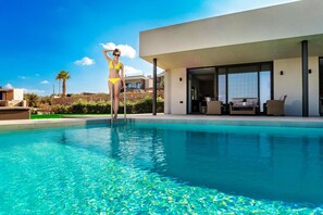 A modern poolside area with a clear blue pool, a person in a yellow swimsuit, and a contemporary building with large windows and outdoor seating. - Villa Kamelia With Pool (Hersonissos)