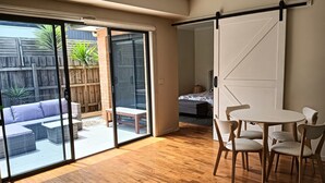 A room with a sliding glass door leading to an outdoor patio, a wooden floor, a round table with chairs, and a bed with a patterned bedspread. - Coastal charm, spacious townhouse Ideal for families or groups of up to 8 (Aspendale)